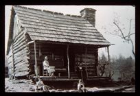 Cabin with three people on porch. Black and white photo. 